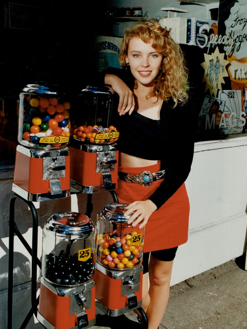 A young Kylie Minogue, wearing black top and red skirt, poses next to a gum ball machine