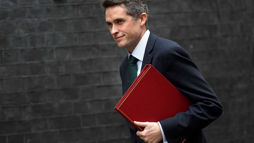 Gavin Williamson holds a red folder as he walks past a brick wall.