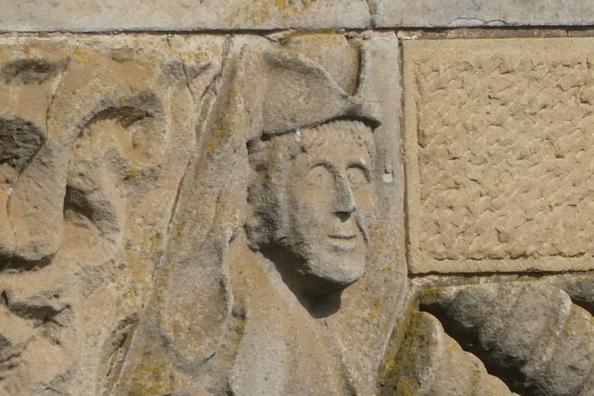 Face carved in bridge in sandstone. Wears hat.