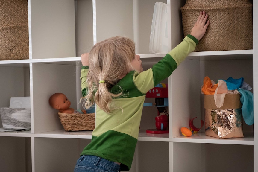 Little girls reaches for a basket on a shelf.