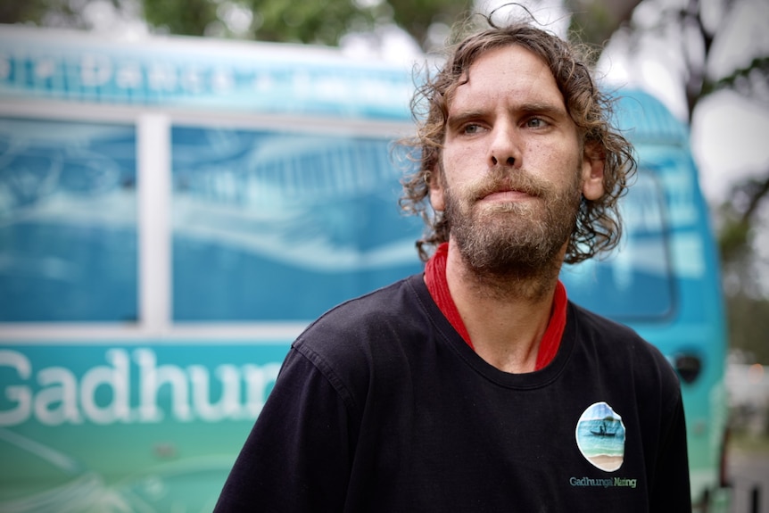 Jacob Morris stands infront of a blue bus wrapped in Aboriginal artwork