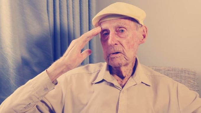 An elderly gentleman with his hand to his face, and wearing a cream-coloured cap and shirt