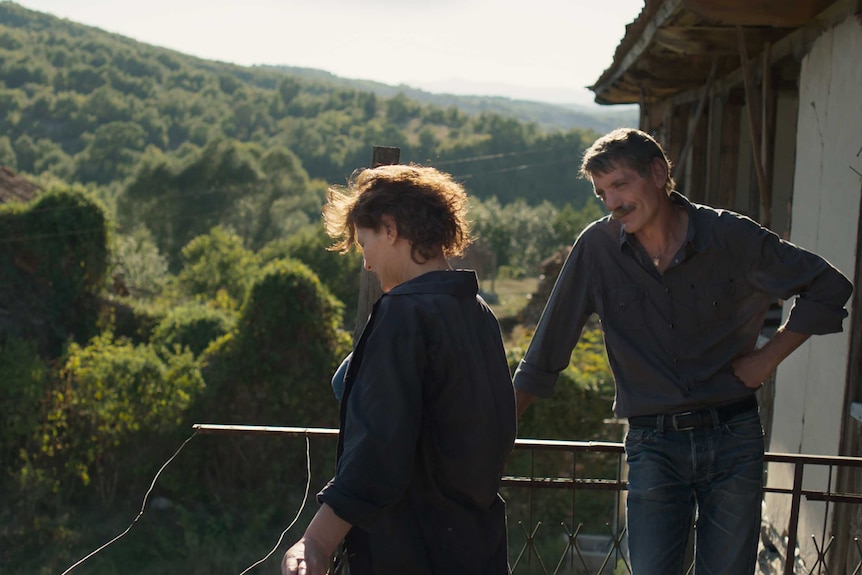 A woman and a man stand talking on a veranda, overlooking a green, treed valley.