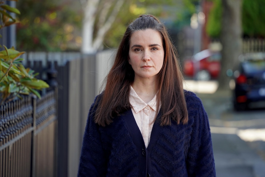 A photo of a woman standing on the footpath of a suburban street, looks straight into the camera with a determined expression.
