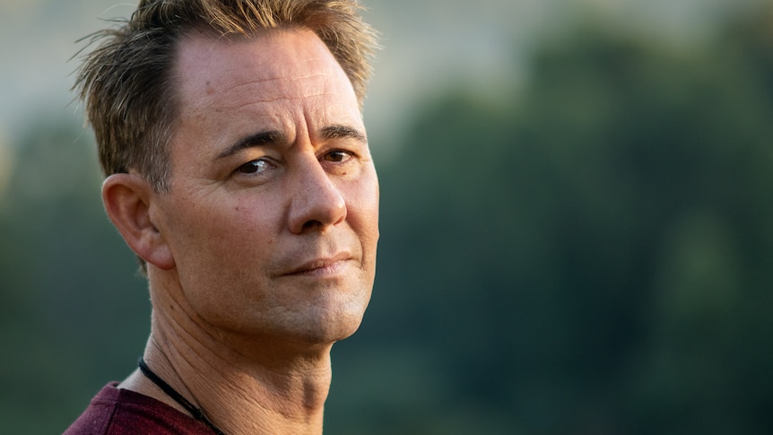 A close up side profile of Jarrod, who looks into the camera with his head tilted, in the late afternoon with green trees behind