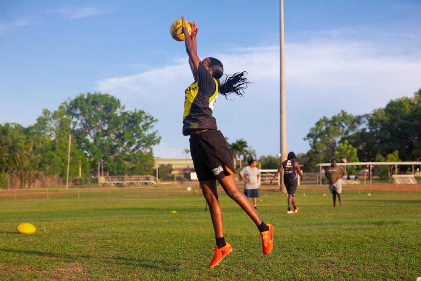 A woman in a black and yellow guernsey marks an Australian Rules football