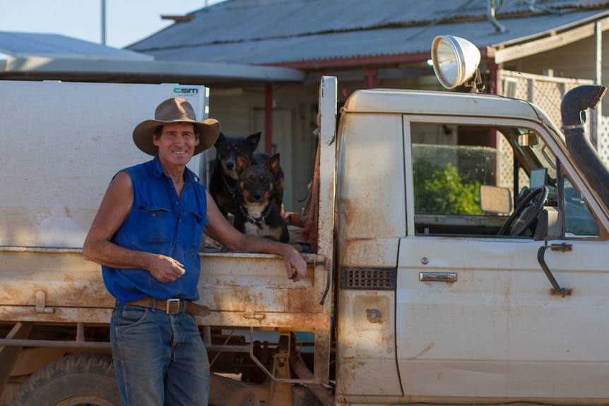 A farmer in a sleeveless shirt and large hat stands by his ute, smiling