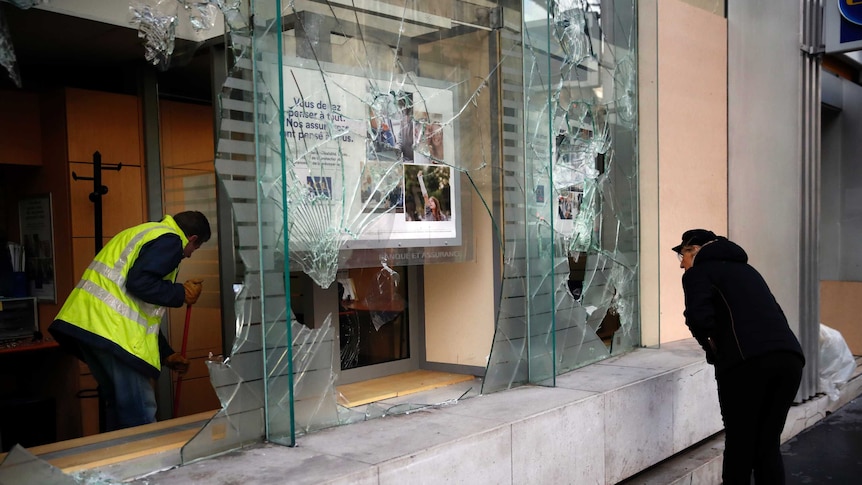 A worker sweeps debris from a smashed glass storefront as a woman looks on.