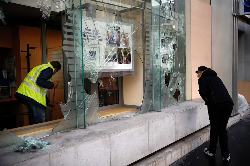 A worker sweeps debris from a smashed glass storefront as a woman looks on.