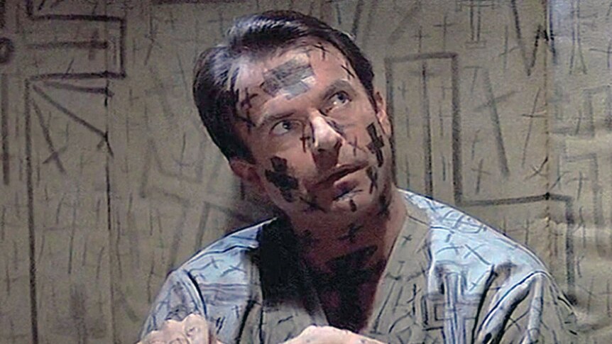 A still from a 90s movie of Sam Neill sitting on the ground, crosses drawn on his faces