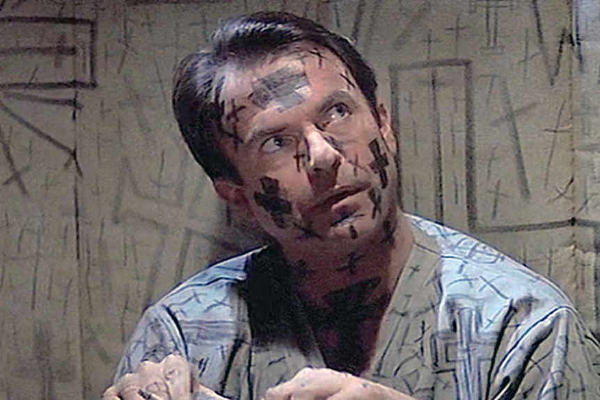 A still from a 90s movie of Sam Neill sitting on the ground, crosses drawn on his faces