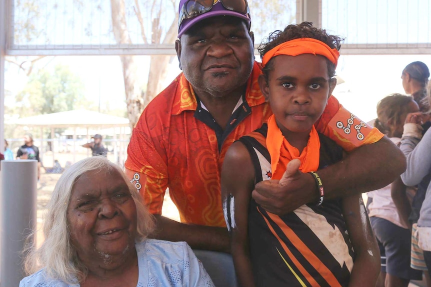 An elderly woman sitting down smiles as a man and a boy stand alongside her smiling.