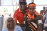 An elderly woman sitting down smiles as a man and a boy stand alongside her smiling.