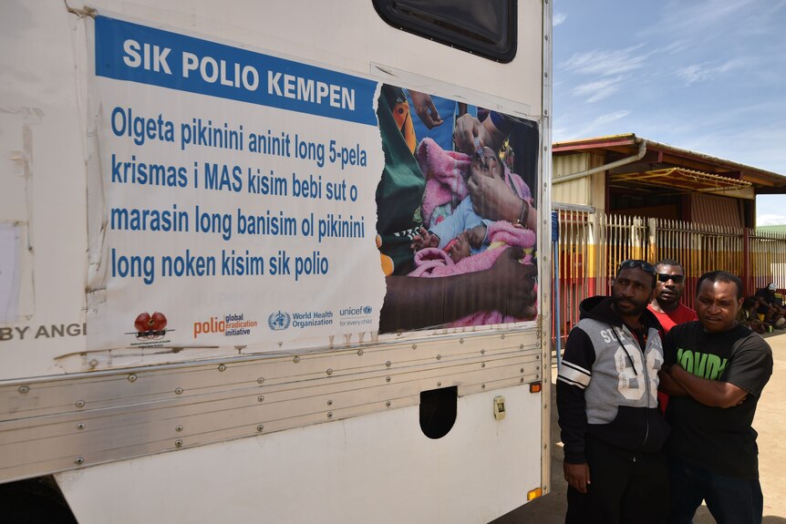 Three men standing to the right of a mobile polio vaccination clinic.