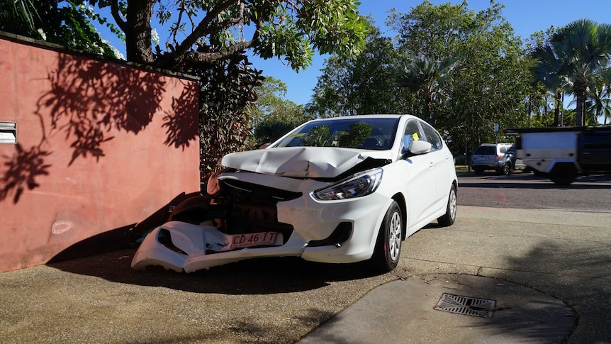 Crashed white car in a driveway in Rapid Creek NT. Sunny day. Trees. Red wall