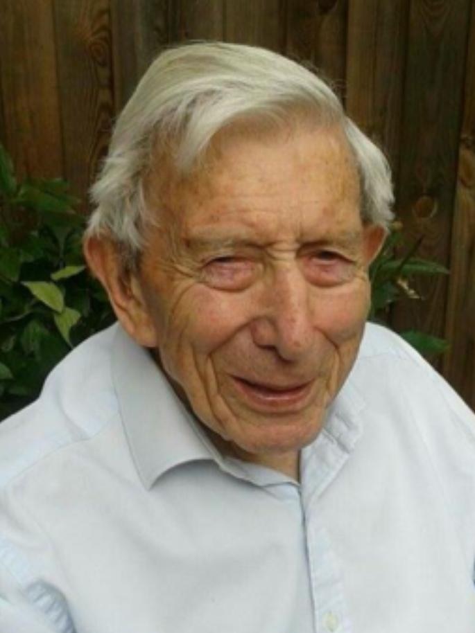 An eldlerly man in a light blue long sleeve shirt, seated and smiling, with a wooden fence behind him