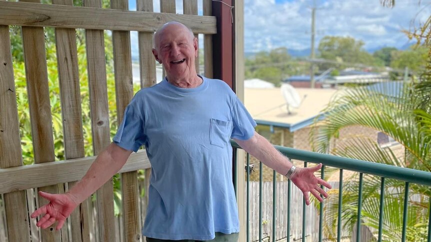 An elderly man with a big smile on his face, standing on the balcony of a unit.