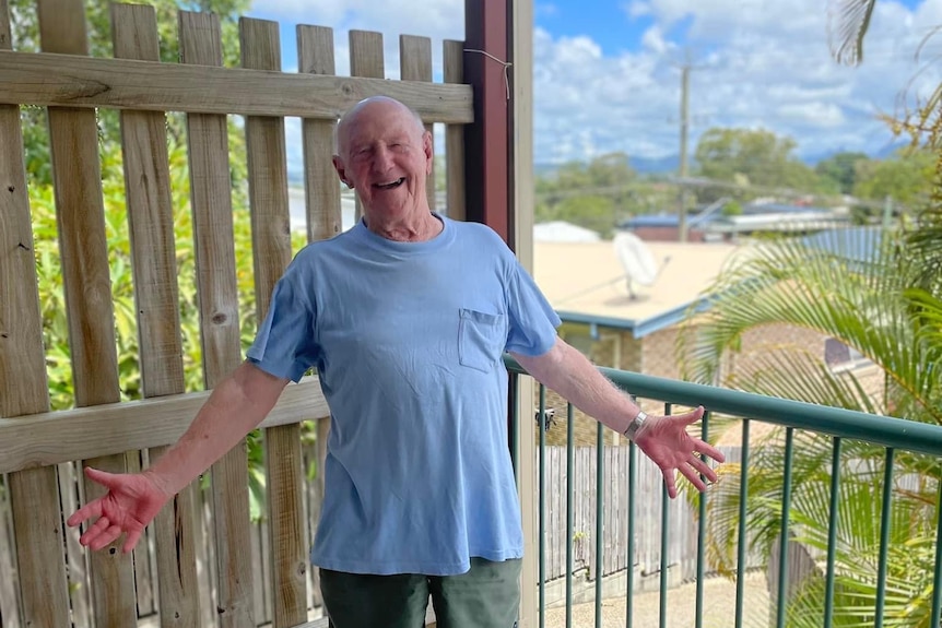 An elderly man with a big smile on his face, standing on the balcony of a unit.