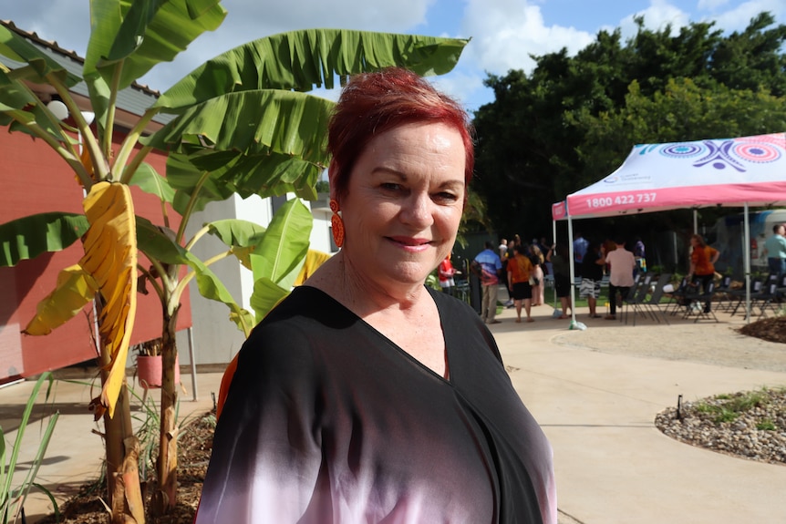 A woman smiling at the camera and standing outside on a sunny day, as a fair happens in the background.