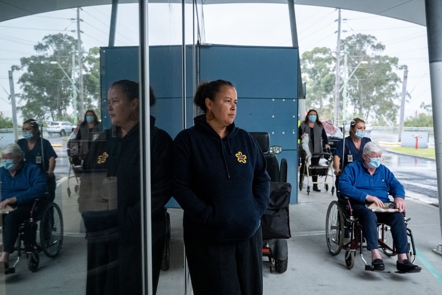 A woman stands outside a hospital. Behind her is an  elderly man in a facemask being pushed in a wheelchair. 