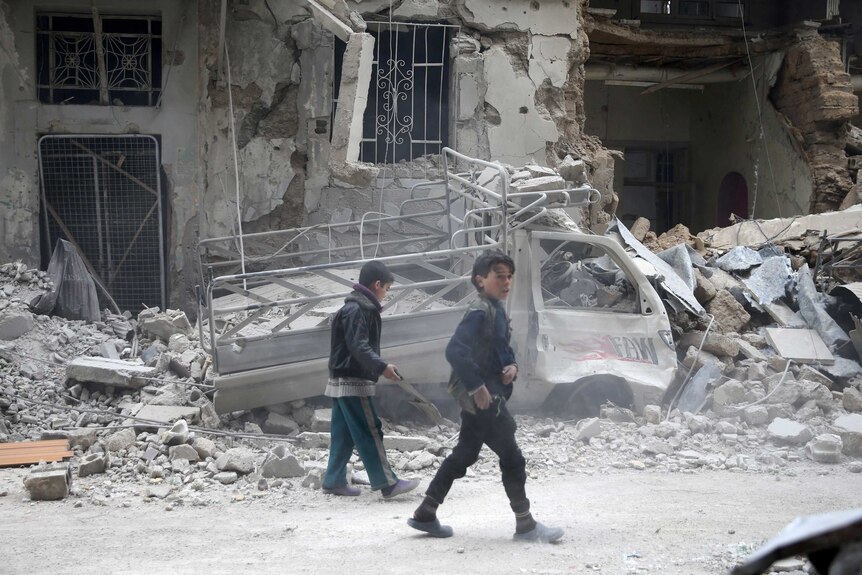 Children walk along a street with rubble an damaged buildings in the background.