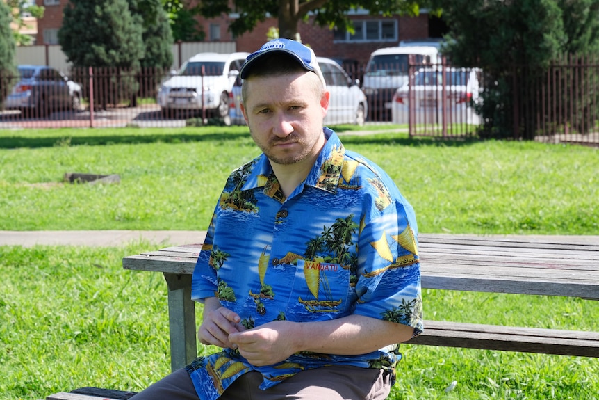  A young man sits on a park bench wearing a blue cap and a Hawaiian shirt.