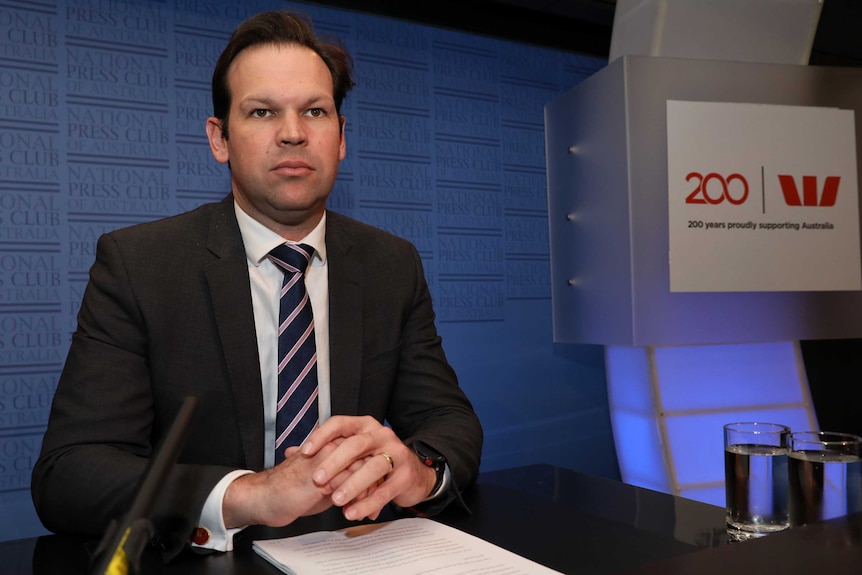 Matt Canavan sits on stage in front of a blue NPC background and stares vacantly past camera.