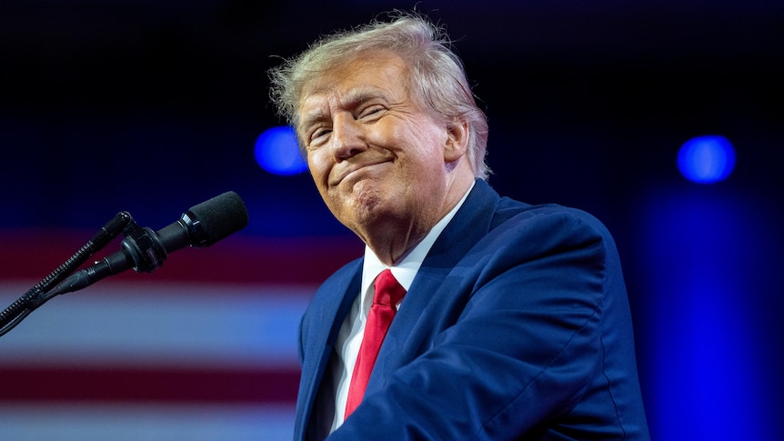 Donald Trump, lit by professional lighting, grins with an American flag visible in the background