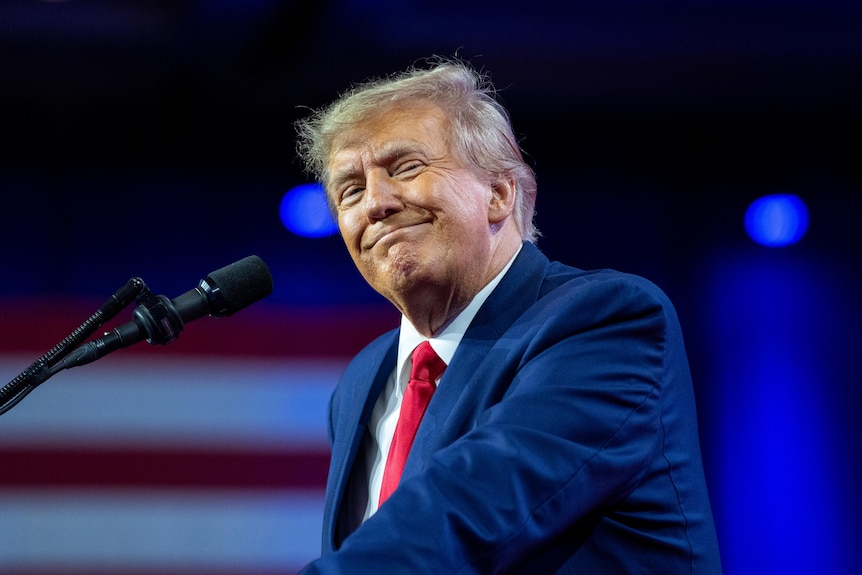 Donald Trump, lit by professional lighting, grins with an American flag visible in the background