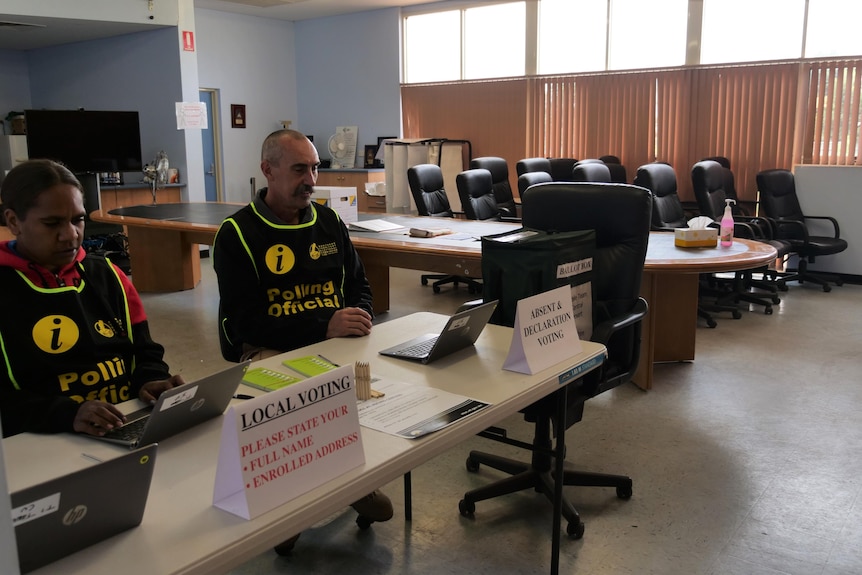 two people wearing polling officer vests sit at a table in an empty room