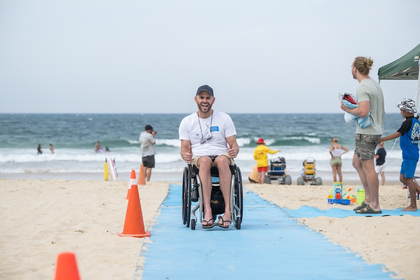 A man laughs and smiles on the beach in a wheelchair on a long blue mat