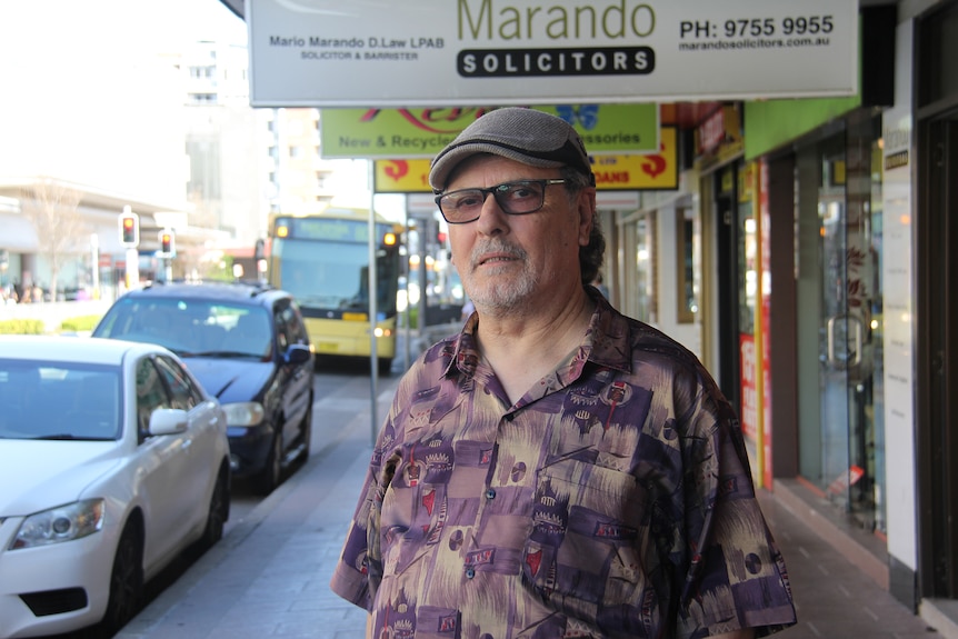 Older man with hat and glasses on standing in street.