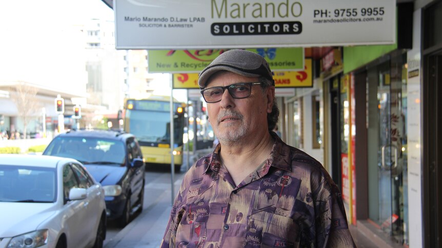 Older man with hat and glasses on standing in street.