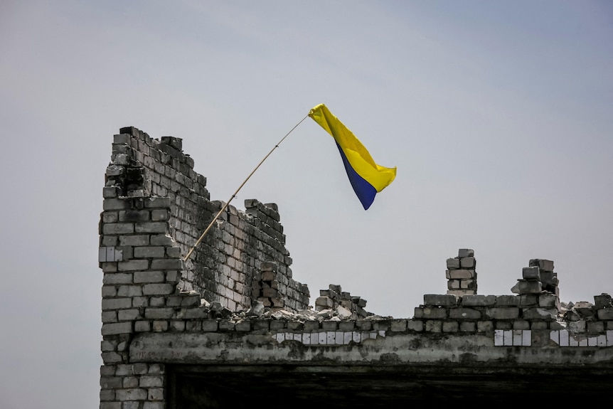 A Ukrainian national flag flutters lightly in a breeze while flying from a makeshift flagpole atop a ruined building.