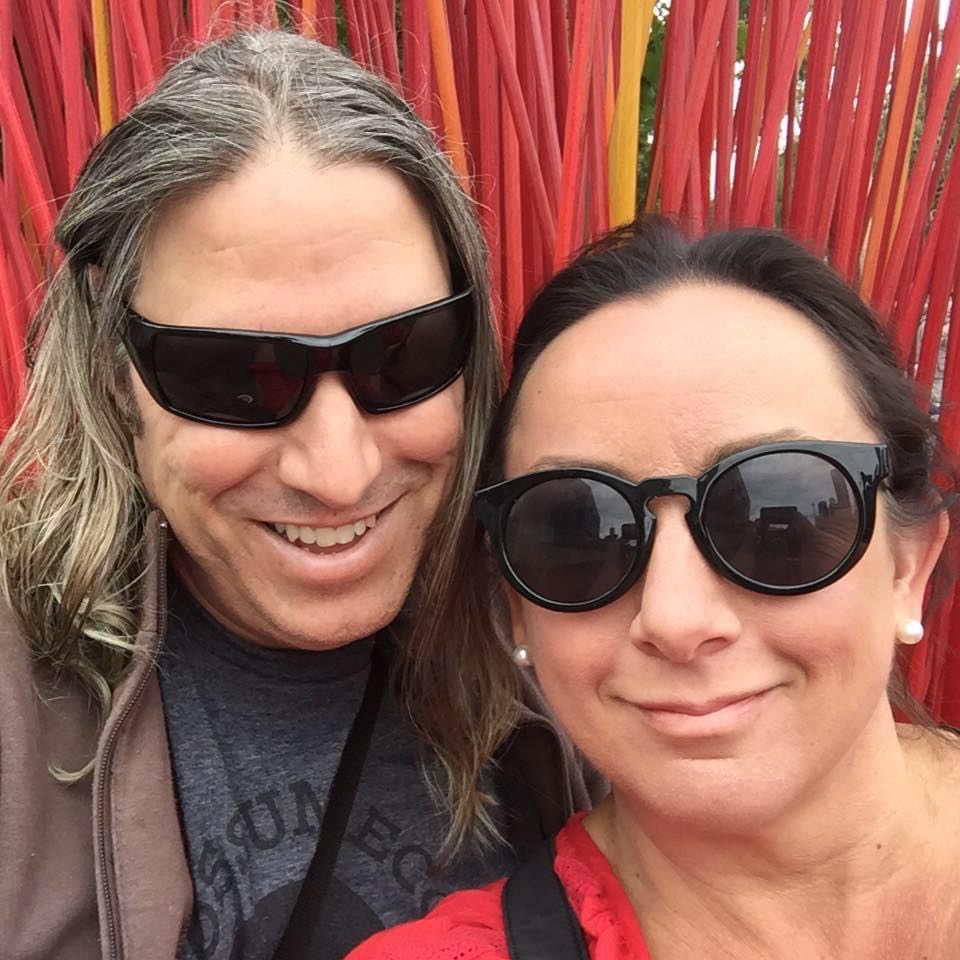 A selfie shot of a man with long gray hair and a woman with dark hair, both wearing sunglasses and smiling.
