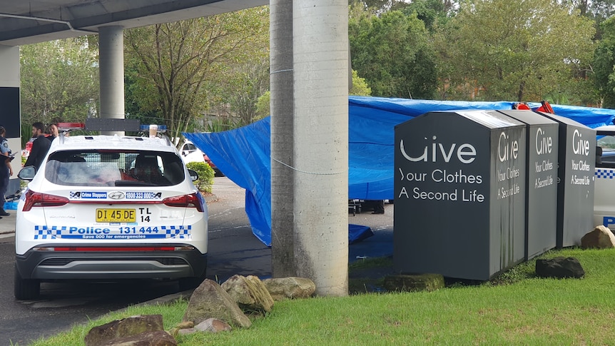 A police car under a bridge next to tarp and three charity bins. 