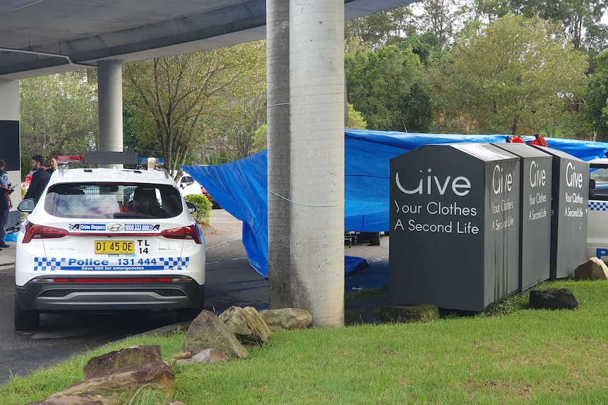 A police car under a bridge next to tarp and three charity bins. 
