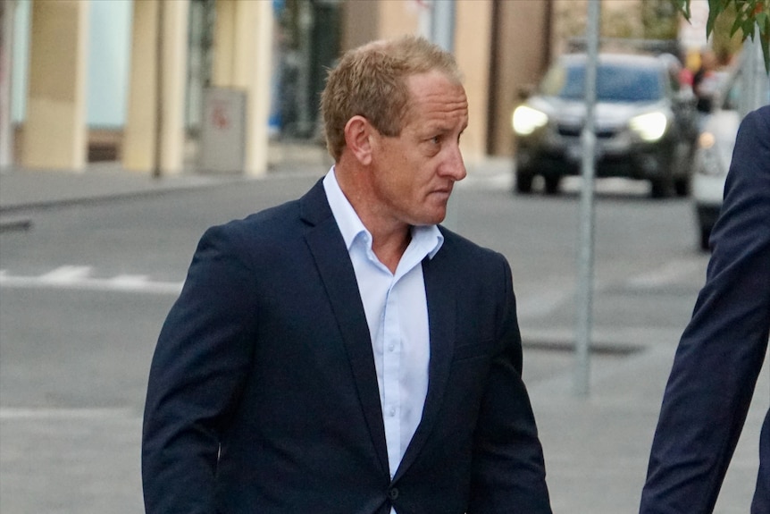 A man with fair hair and skin, wearing a suit jacket and an open-necked shirt.