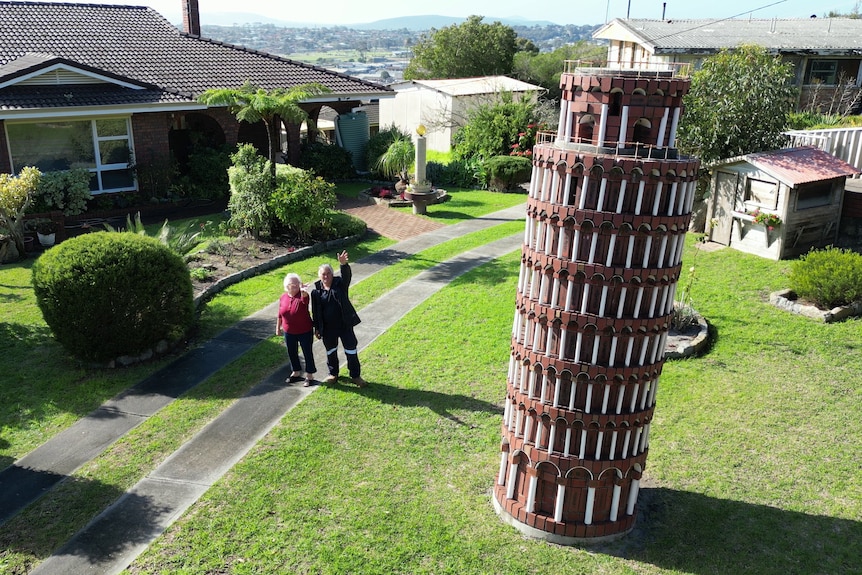 Two people wave in a front yard next to a leaning tower. 