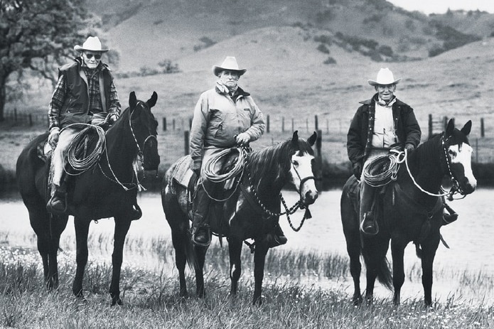 Three cowboys on their horses in a field with a small hill in the background in black and white.