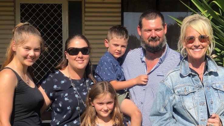 Three adults and three children casually dressed stand close together smiling and squinting into the sun.