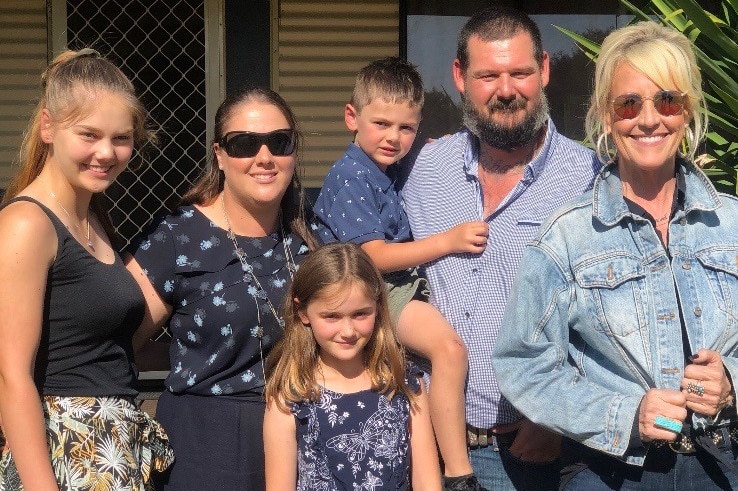 Three adults and three children casually dressed stand close together smiling and squinting into the sun.