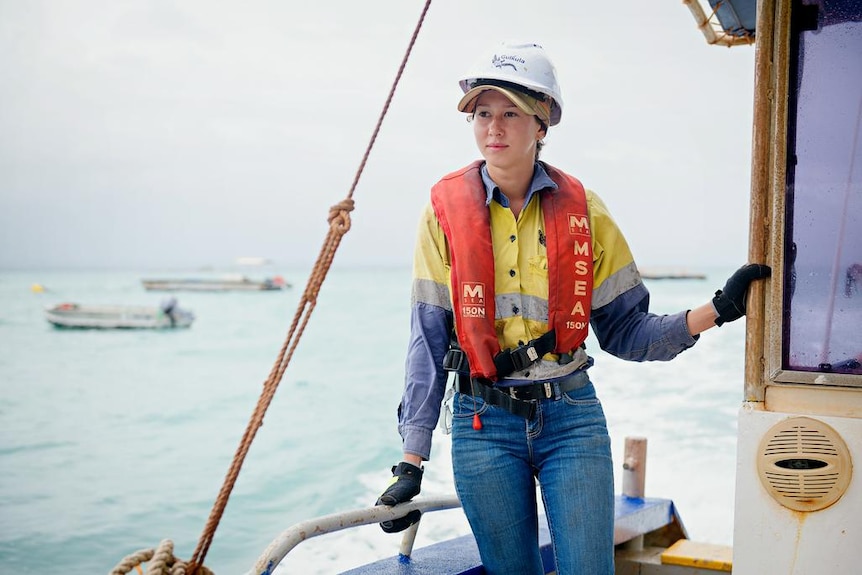 A young woman hanging on to some railings on a fishing-type boat, wearing a high-vis shirt, helmet and life jacket.