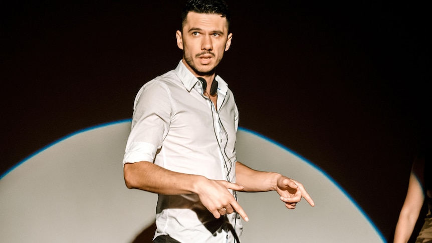 Goran Stolevski stands in a spotlight on a stage, wearing a white button up and blue jeans.