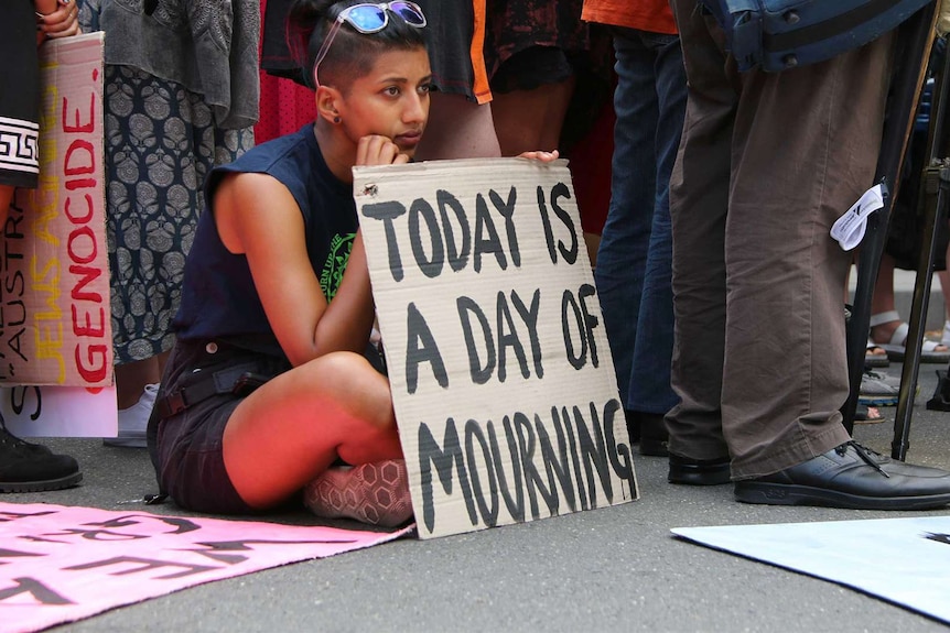 A woman sits on the pavement. Her cardboard sign reads "Today is a day of mourning".