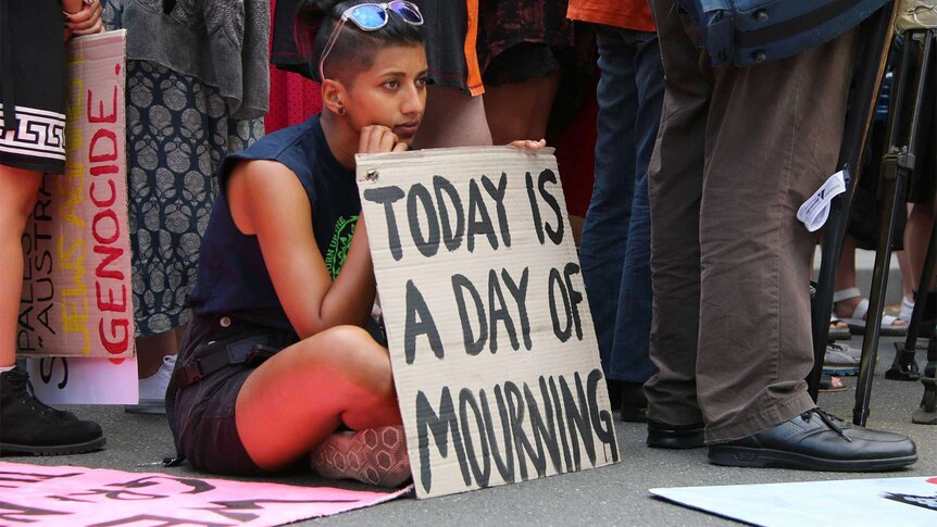 A woman sits on the pavement. Her cardboard sign reads "Today is a day of mourning".