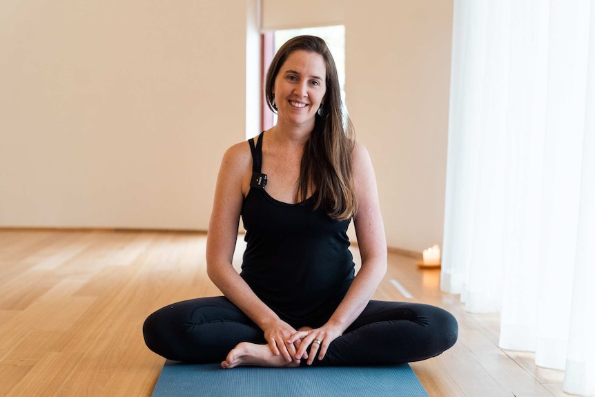 A woman with long dark hair sits on a yoga mat smiling with her legs crossed wearing black.