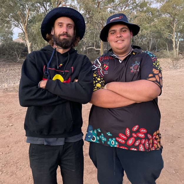 two aboriginal men arms crossed in front of them wearing black on red soil with trees in background