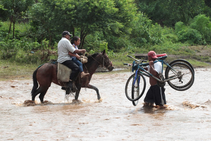 A man and child on a horse pass a man carrying two bicycles as they cross a flooded river.