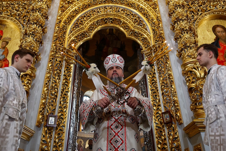 An orthodox priest conducts a ceremony in a gilded church with two men in silver robes standing each side of him